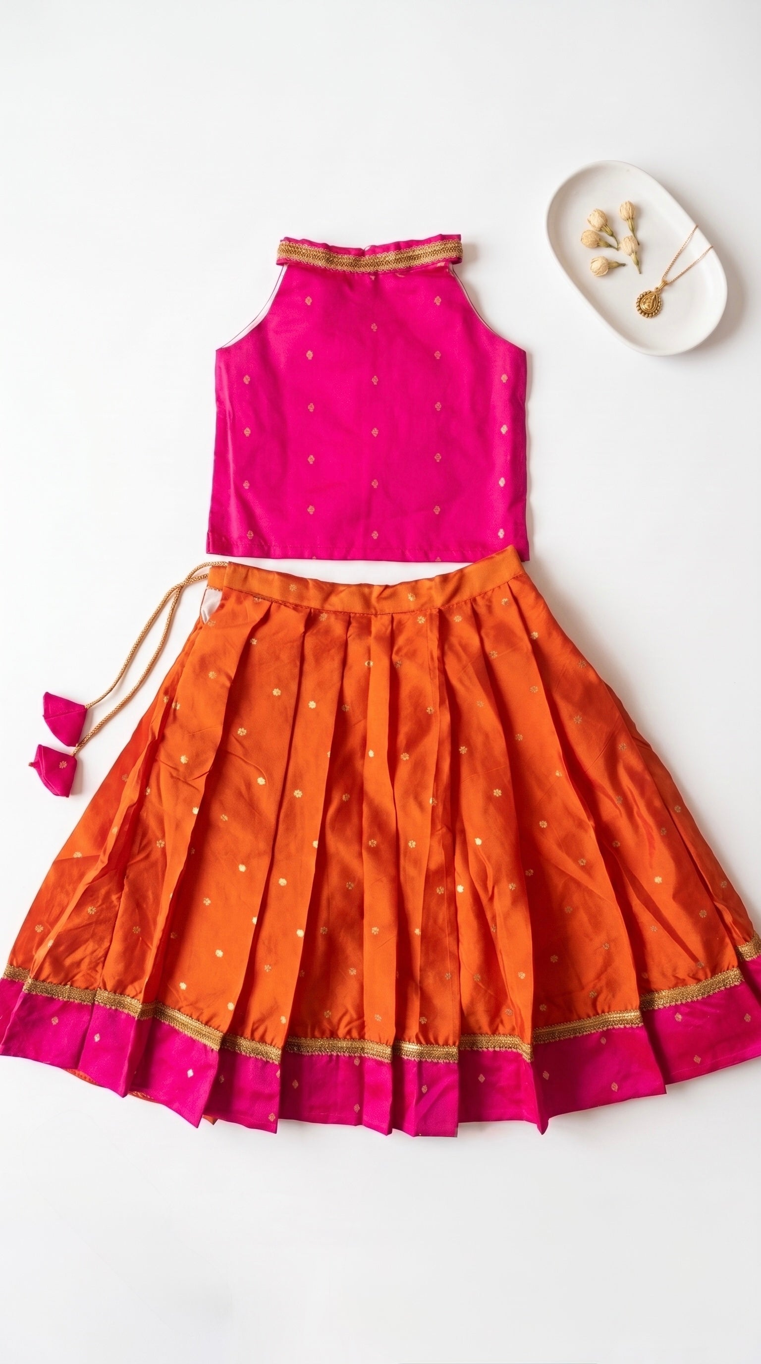 A two-piece children's traditional outfit laid flat on a white background, featuring a sleeveless bright pink top with gold trim and a matching pleated orange skirt with gold polka dots and a pink border.